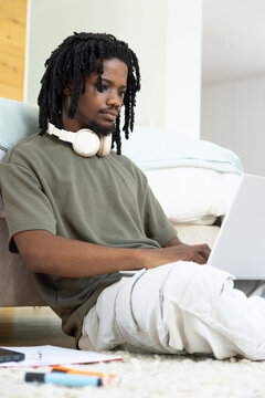 Man with dreadlocks wearing olive tee sitting on carpet using laptop, white headphones nearby
