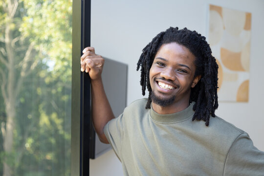African American man leaning on sliding door frame at home, smiling in green sweatshirt, art behind