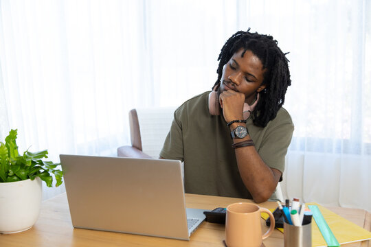 Adult African American man sitting at home desk, leaning on fist, using laptop with pink headphones