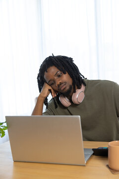 African man sitting at table at home, leaning on hand, viewing silver laptop with pink headphones