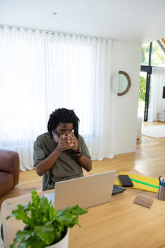 Man sitting at table using silver laptop in living room holding mug wearing olive tee headphones