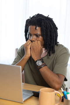 African American man sitting at wooden table at home, leaning on fists, studying laptop and mug