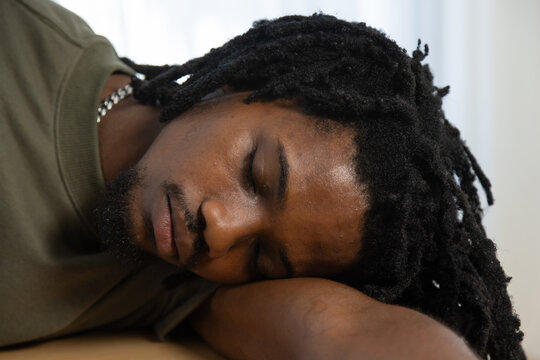 African American man resting with eyes closed on table at home, wearing olive T-shirt, silver chain