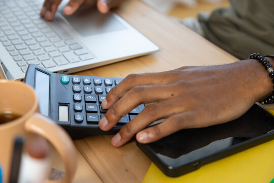 African American man pressing calculator buttons using laptop at desk with mug and beaded bracelet