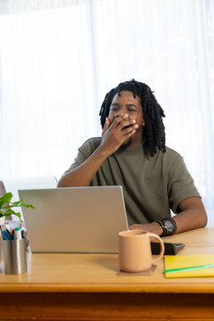 African man in his 20s yawning, covering mouth, working at wooden desk with laptop, mug, plant