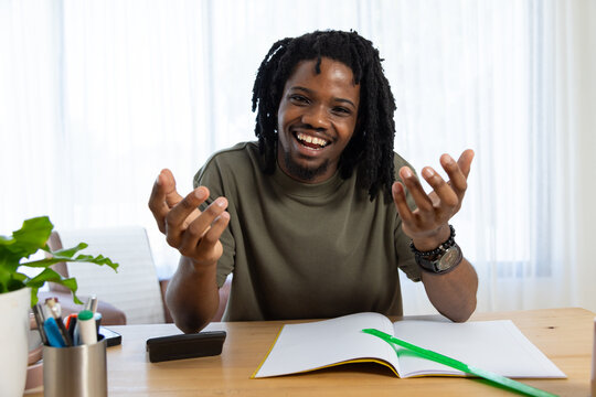 Adult African man smiling, sitting at wooden table in bright room with notebook, green pen, device