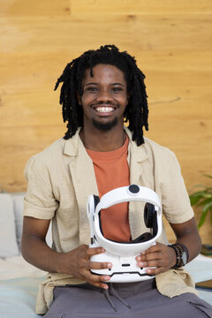 African American man smiling and sitting at home against wooden panel wall holding white VR headset
