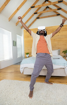 Man wearing VR headset and headphones standing barefoot on rug in modern bedroom raising arms