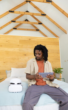African American man sitting on bed holding tablet in loft bedroom beside VR headset