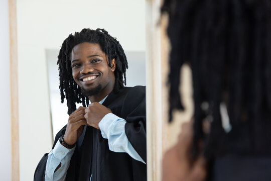 Adult African American man adjusting black academic gown, checking mirror at home