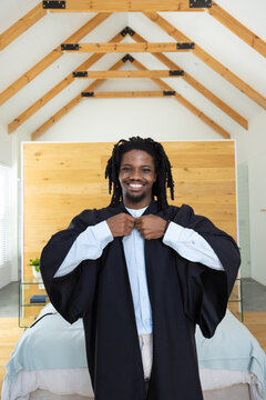 Smiling man adjusting black graduation gown while standing in bedroom with wooden headboard and bed