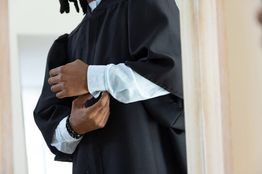 African male adjusting shirt cuff and arranging black gown near mirror with beaded bracelet