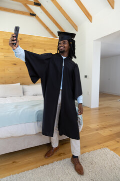 African American man wearing black gown and mortarboard holding smartphone, smiling in bedroom