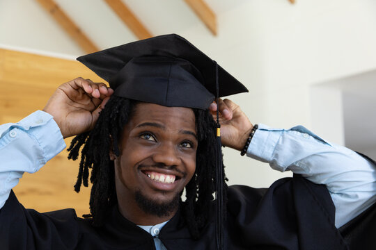 African American adult man adjusting mortarboard and gown, smiling upward in wood-paneled room
