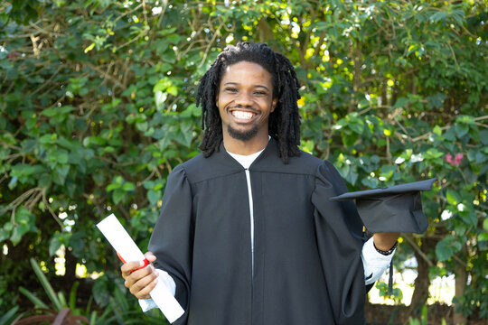 Man smiling and holding red-tied diploma and cap, wearing gown and bracelet in garden