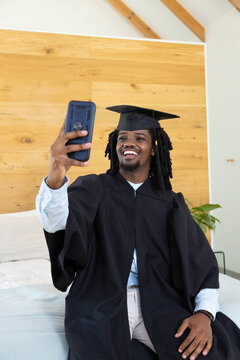 African American man in 20s wearing black gown and cap, taking selfie in bedroom with smartphone