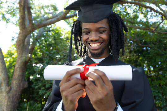 African American man wearing mortarboard and gown, holding rolled diploma with red ribbon outdoors