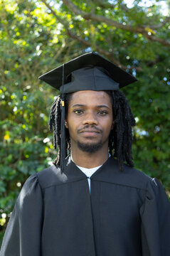 African American male wearing mortarboard and gown standing in sunlit park with tassel visible