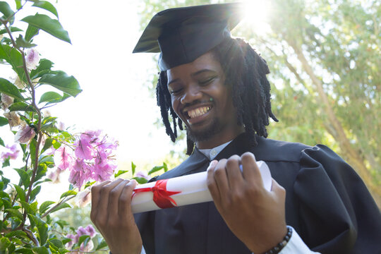 African American man standing in garden wearing mortarboard and gown, holding rolled diploma