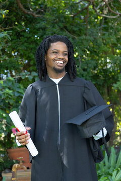 African man wearing gown standing in garden holding cap and rolled diploma by planter and tap