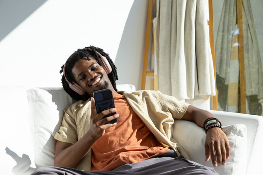 Adult male reclining on sofa in living room, wearing pink headphones and holding smartphone