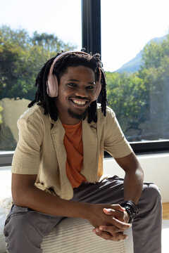 African man sitting on cushioned seat by large window wearing pink headphones and bracelets
