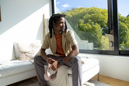 Adult African man sitting on chaise in living room wearing rust T-shirt holding pink headphones