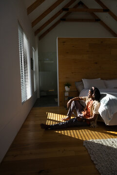 Adult African man sitting on wood floor next to bed holding smartphone, headphones on, copy space