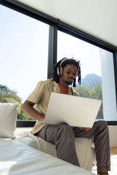 African American man sitting on window seat in bright living area, wearing headphones, using laptop