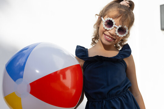 Female child standing against sunlit white wall in navy dress holding bright beach ball, copy space