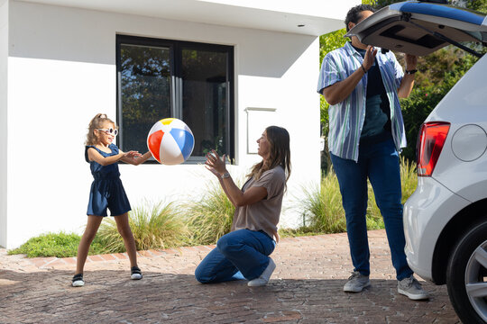 Diverse family playing on driveway while child girl with shades tossing beach ball near car trunk