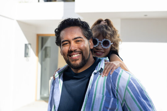 African American dad carrying daughter on back at white home wearing striped shirt, flower shades