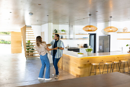 Couple dancing and holding hands at home in modern kitchen near wooden island and stools