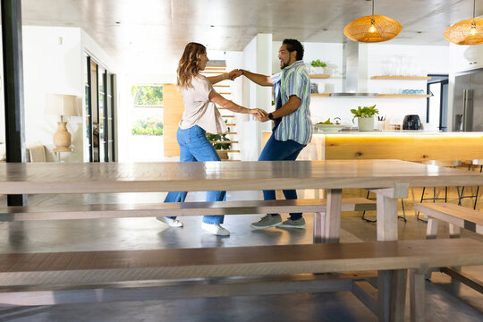 Couple in casual clothing dancing and holding hands in open kitchen near wooden dining table