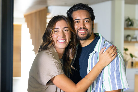 Diverse couple smiling and embracing while standing in modern kitchen near wooden slat divider