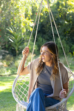 Woman sitting in woven swing 3 ropes fringe wearing jeans jewelry smiling looking right in garden