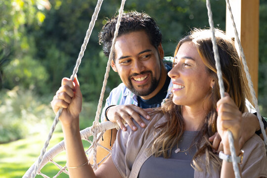 Couple sitting on woven rope swing holding thick ropes in sunny backyard, showing pendant necklace
