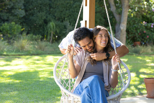 Couple sharing affectionate hug while sitting in macrame swing with ropes and watches in backyard