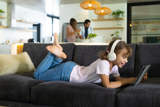 Diverse family relaxing in living room, child lying on sofa using tablet with white headphones