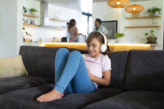 Family with child wearing pink top and white headphones sitting on gray sofa using tablet