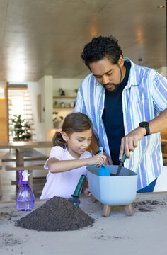 African American dad and girl potting soil in planter at table using trowel and blue scoop