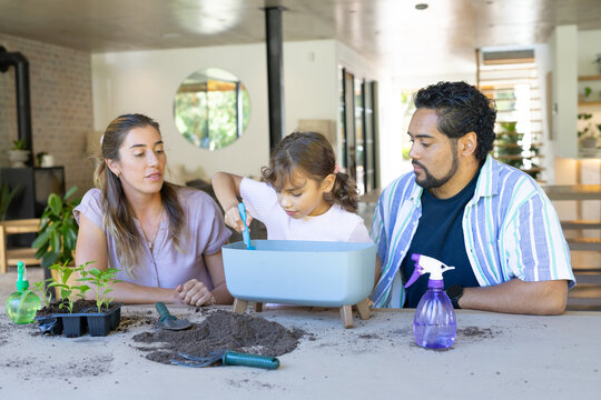 Diverse family parents and daughter potting plants at home table with light-blue planter and trowel