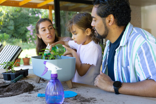 Parents and daughter potting plants on covered patio, child watering gray planter with green bottle