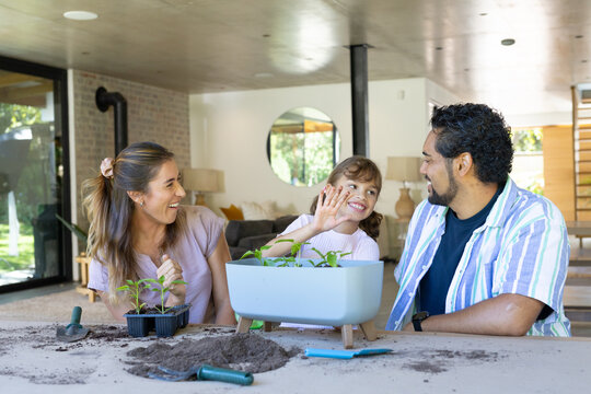 Diverse family with child planting seedlings at dining table in living room with planter and trowel