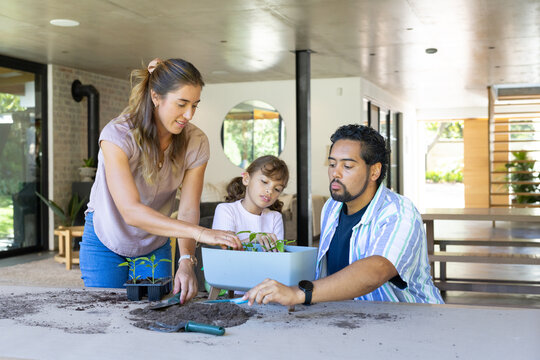 Diverse family with child potting seedlings at home around planter, scooping soil with trowel