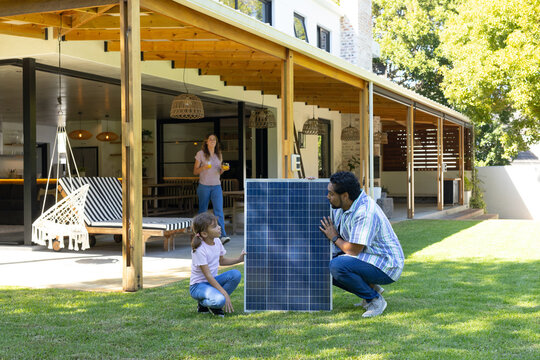 Diverse family kneeling by solar panel in sunny backyard under wooden pergola, woman holding cup