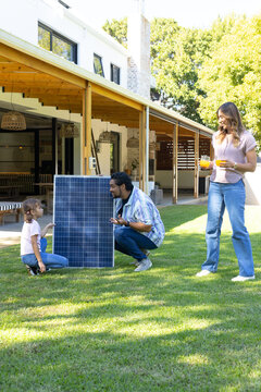 Family with child crouching and kneeling in backyard near solar panel, woman holding two drinks