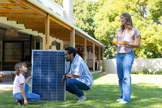 Diverse family crouching by solar panel outdoors, child pointing, woman holding two orange juices