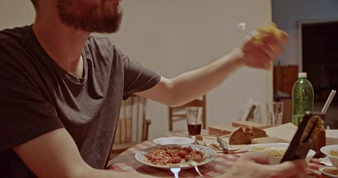 A young man with a beard sits at a table, enjoying his dinner. He is casually using his smartphone, highlighting the common modern habit of digital interaction during meals.