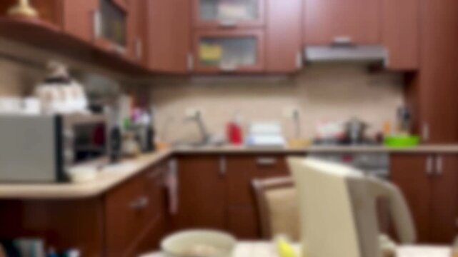 Blurred kitchen interior warm wooden cabinets, soft focus view of counter with microwave, sink, chair and stool, kettle on tile